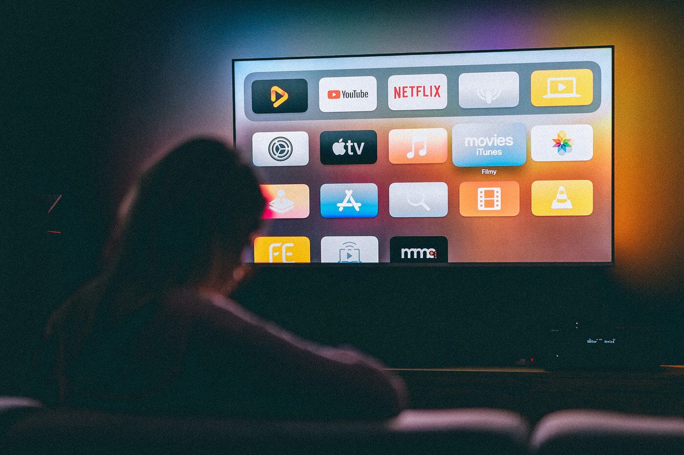 A person sits in a dimly lit room, facing a large screen displaying various app icons including YouTube, Netflix, Apple TV, and others. The room has a dark ambiance, and the screen glows brightly with vibrant colors from the app icons. IPTVMAX